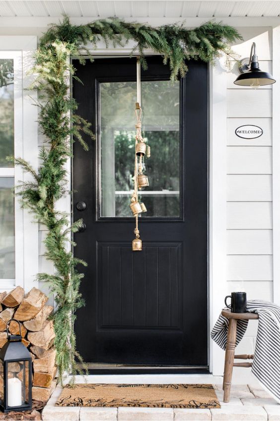 a simple rustic front porch with a fir garland, firewood, a candle lantern, bells and a wooden stool with a blanket