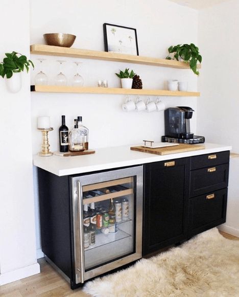 A small and catchy home bar with light stained open shelves, black cabinetry, a wine cooler, various plants, bottles and candles