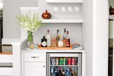 a small and lovely built-in home bar with a white cabinet, a wine cooler, open shelves, blooms, beautiful vases and glasses and wine bottles