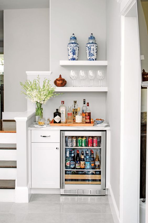 a small and lovely built-in home bar with a white cabinet, a wine cooler, open shelves, blooms, beautiful vases and glasses and wine bottles
