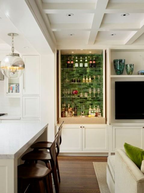 A small built in home bar with green tiles, glass shelves, built in lights, a cabinet and a sink plus lots of glasses and wine bottles