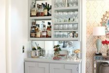 a stylish built-in home bar in dove grey, with a mirror backsplash, open storage compartments and a cabinet plus plants and blooms