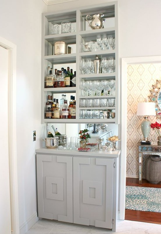 A stylish built in home bar in dove grey, with a mirror backsplash, open storage compartments and a cabinet plus plants and blooms