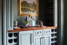 a vintage built-in home bar with blue cabinetry, wine storage compartments, artwork, wine bottles and wine glasses