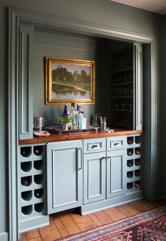 A vintage built in home bar with blue cabinetry, wine storage compartments, artwork, wine bottles and wine glasses