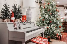 a vintage grey piano with some Christmas trees, a gnome, a grey bench with a printed red blanket, a Christmas tree with red and white ornaments