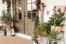 a vintage rustic front porch with mini trees and fir branches in baskets and buckets, candle lanterns, a fir garland, red touches