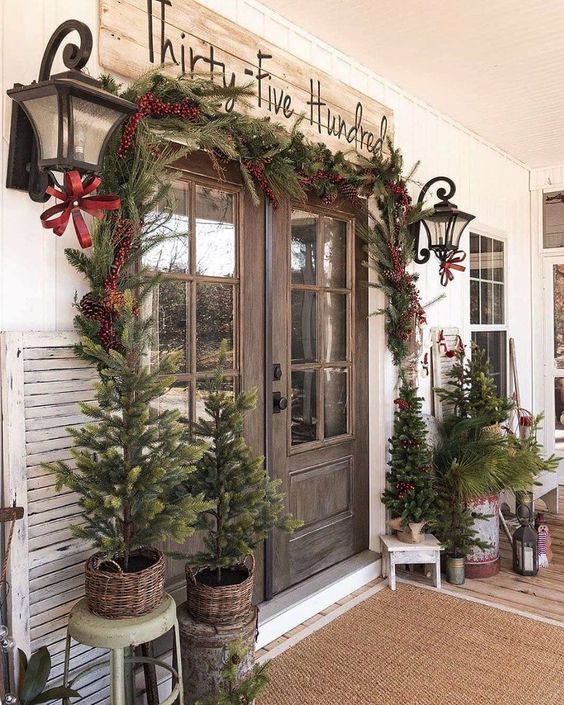 a vintage rustic porch with lots of Christmas trees in baskets, fir branches in a a bucket, lanterns, a fir garland with berries around the door