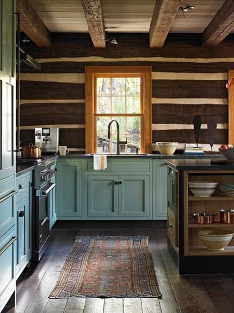 a welcoming cottage kitchen with wooden beams on the ceiling, dark stained planks on the walls, blue shaker style cabinets and a small kitchen island with open storage compartments