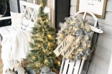 elegant white farmhouse Christmas decor with a white sleigh with a flocked wreath and lights, a white lantern, a mini tree with lights, a bucket with branches and a white bench with a blanket