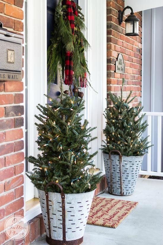 mini Christmas trees in buckets with lights, a fir branch hanging with a plaid scarf make the porch look rustic and cozy