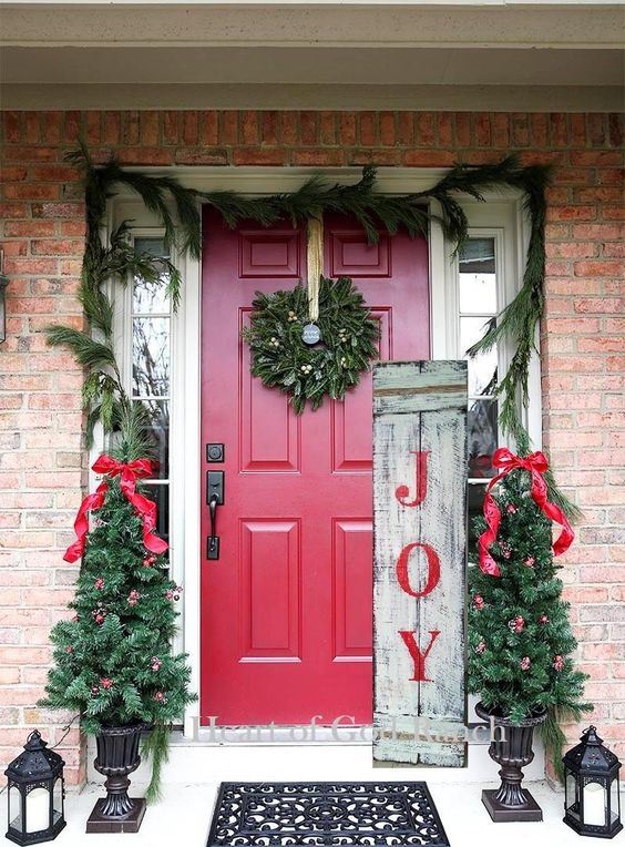 rustic Christmas front porch decor with an evergreen garland, mini trees with red ornaments and bows, a sign and candle lanterns