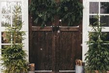 simple rustic front porch decor with Christmas trees in crates, buckets with firewood, fir wreaths on ribbons looks al-natural