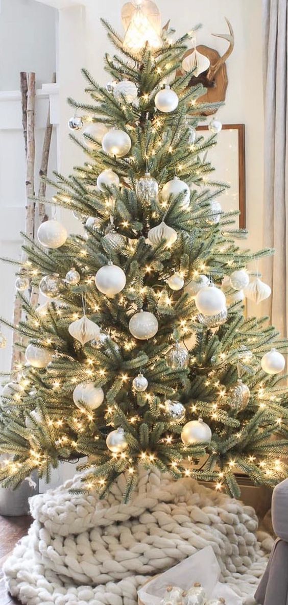 a Christmas tree with oversized white and silver ornaments and lights and a white chunky knit tree base cover is cool