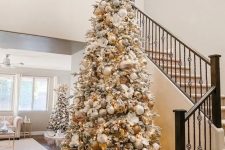 a flocked Christmas tree with white, silver and gold ornaments, white faux blooms and gold leaves and branches on top