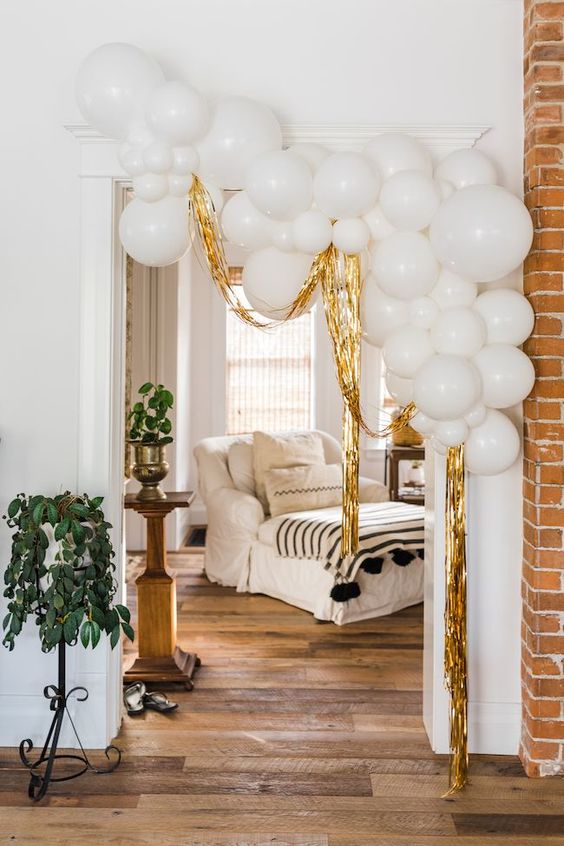 A lovely and modern NYE party decoration   an arrangement of white balloons with gold tinsel is a cool decor idea