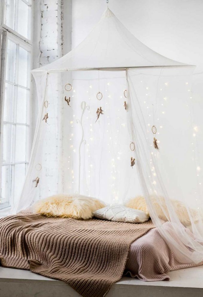 a neutral bedroom with a low bed right on the floor, pastel bedding, a semi sheer canopy with lights and dream catchers over the bed