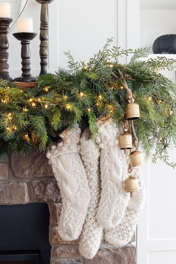 a pretty woodland or cabin mantel with an evergreen and light garland, vintage bells and white knit stockings for Christmas