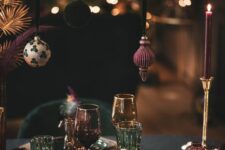 a sophsticated NYE party tablescape in jewel tones, with Christmas ornaments hanging over the table, gold placemats, gold candleholders and purple candles