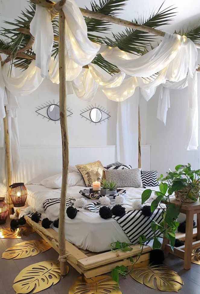 A tropical inspired bedroom wih a pallet and branch bed, black and white bedding, potted greenery, candle lanterns and curtains with lights over the bed