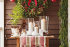a console table with pillar candles, pinecones, an evergreen wreath with red berries and bows, red lanterns and firewood in a bucket