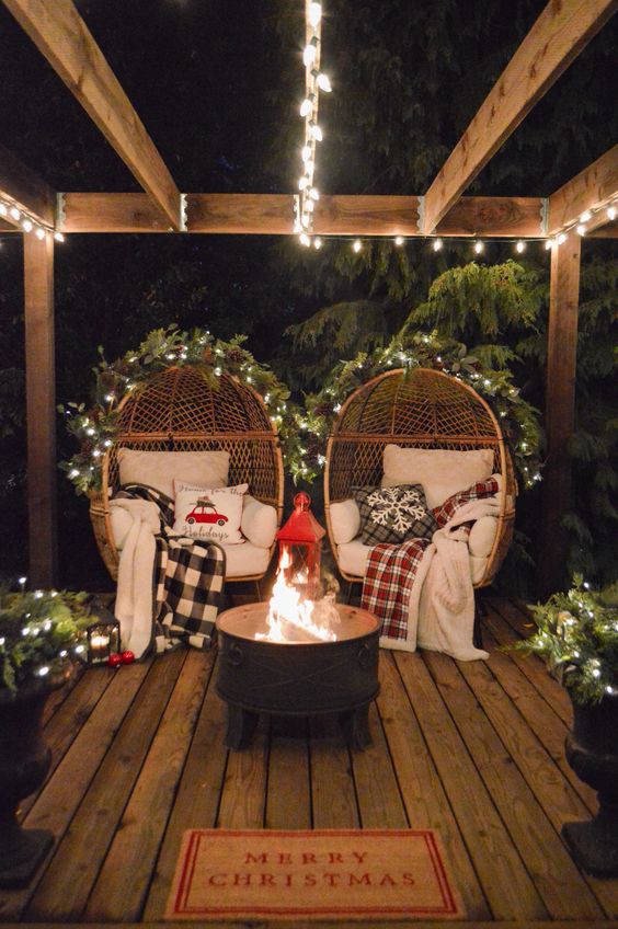 A cozy Christmas decor with egg shaped chairs with lots of pillows and blankets, a hearth, a lantern, lights and urns with lights and evergreens