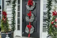 a festive Christmas porch with an evergreen garland with lights and red ribbons, snowy wreaths with red bows and mini Christmas trees