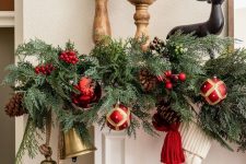 a lush and bright Christmas garland of evergreens, red ornaments, pinecones, berries and large bells is a cool decor idea for the holidays