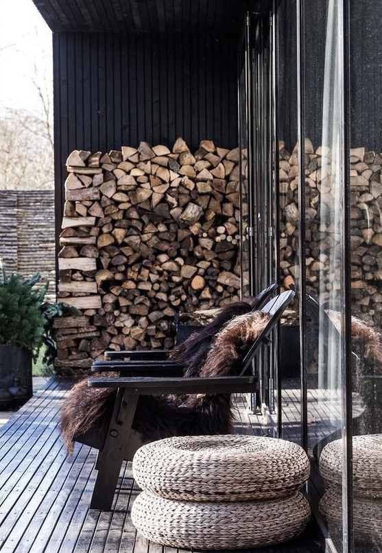 a small and cozy winter deck with black wooden chairs and faux fur, firewood and jute poufs and greenery in pots