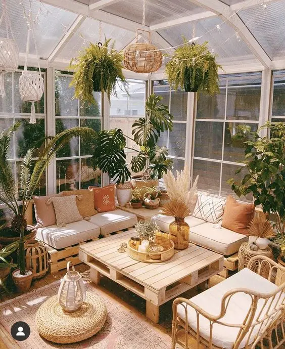 a beautiful boho sunroom with a pallet sectional sofa, a pallet table, a rattan chair, potted plants all over, pendant lamps and a jute pouf