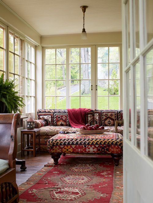 A bold boho sunroom with a red printed boho sofa, a matching ottoman, a red printed rug, rich stained furniture and potted greenery