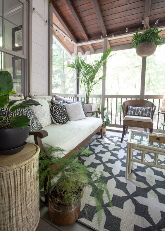 a lovely boho sunroom with a printed rug and pillows, a white sofa, wicker chairs and a rattan table plus potted greenery