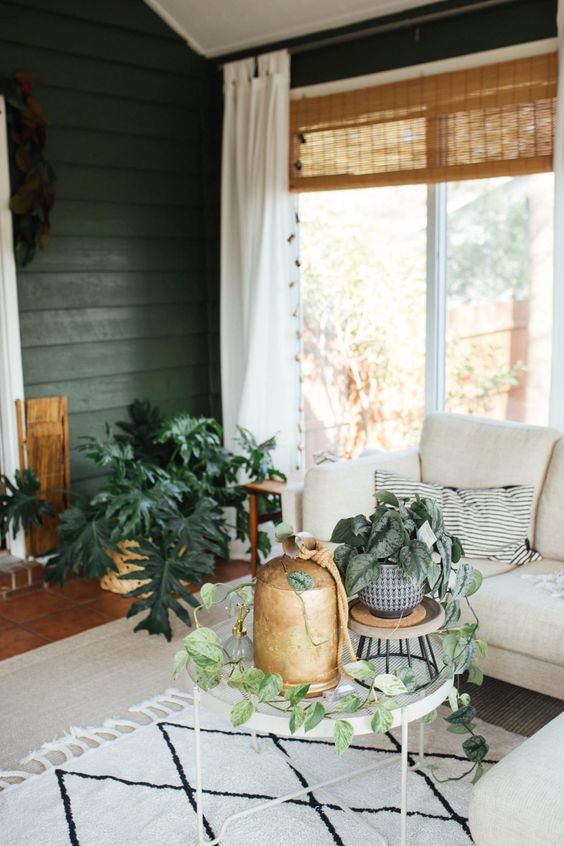 a mid-century modern boho sunroom with green walls, a neutral sectional, round tables, potted plants and woven shades