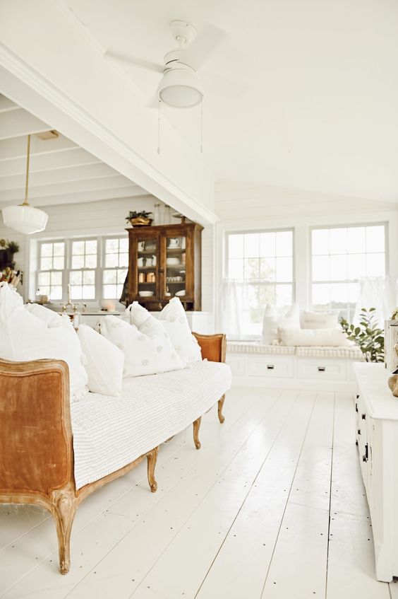 a French farmhouse sunroom done in white with an L-shaped white window seat, a refined sofa and potted greenery