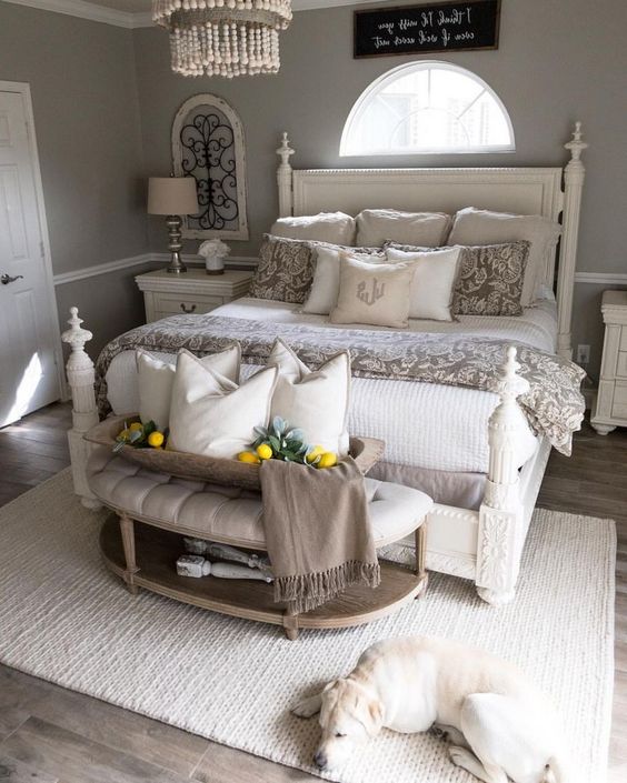 a chic taupe and creamy farmhouse bedroom with elegant furniture, a bead chandelier and an uphostered ottoman