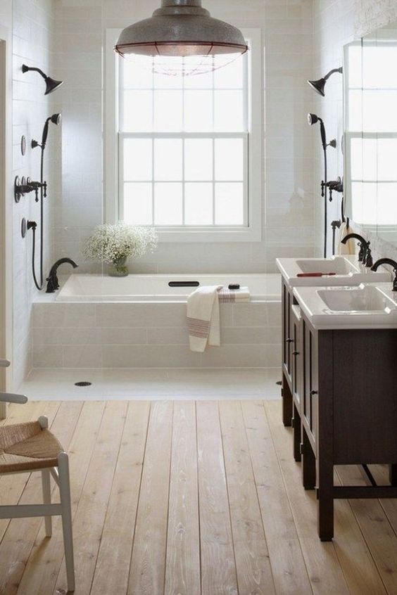 a light-filled farmhouse bathroom with a wooden floor, white tiles, dark wooden vanities and black fixtures