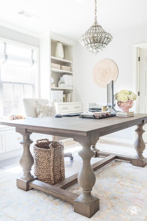 A light filled farmhouse home office with a large wooden desk, neutral storage units, a crystal chandelier and neutral blooms