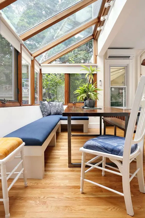 A modern sunroom with a built in bench with navy upholstery, a dark stained table and white chairs and a glazed roof