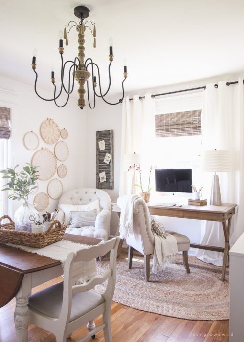 a neutral and cozy farmhouse home office with a wooden desk and a table, neutral upholstered chairs, a vintage chandelier, macrame art on the wall