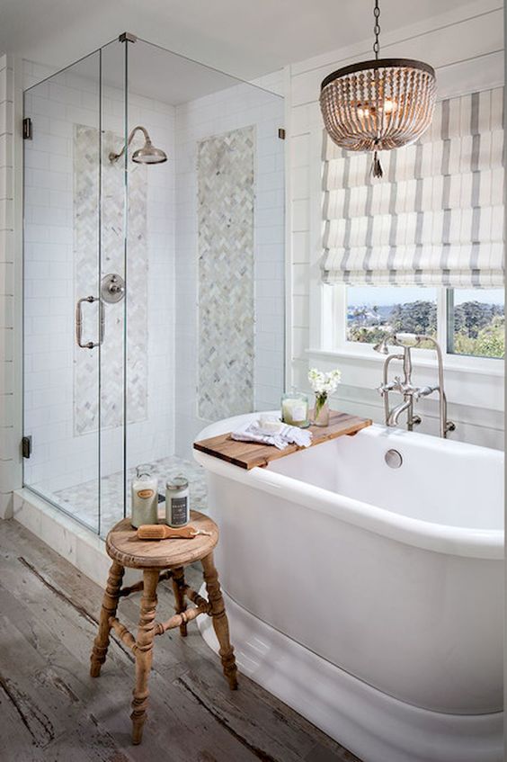 a neutral farmhouse bathroom with marble tiles, Roman shades, a bead chandelier and a vintage wooden stool