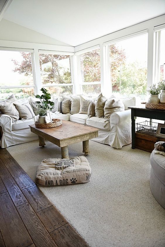 A simple farmhouse sunroom with an L shaped white sofa, potted greenery, a rustic table and neutral textiles