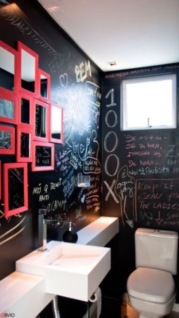 a small modern powder room with black chalkboard walls, a white wall-mounted sink, a white toilet and a gallery wall of pink framed mirrors