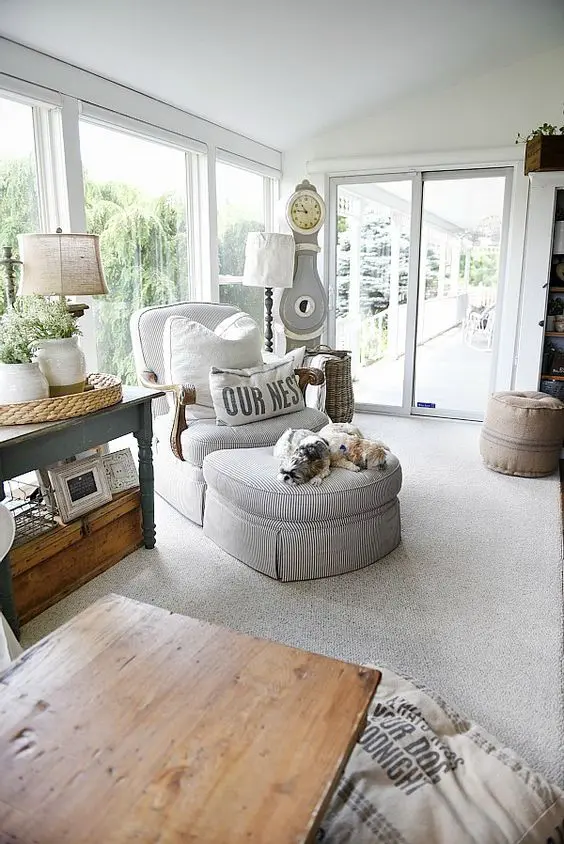 a vintage farmhouse sunroom with vintage rustic furniture, a striped lounger, lamps, greenery and a clock in neutrals