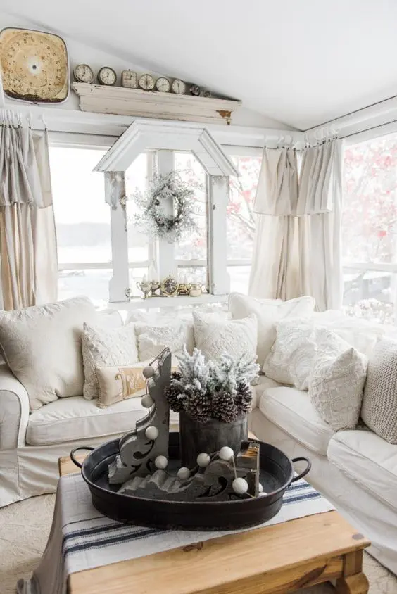 a welcoming neutral farmhouse sunroom with a sectional, vintage decor, wreaths, clocks and textiles in creamy shades