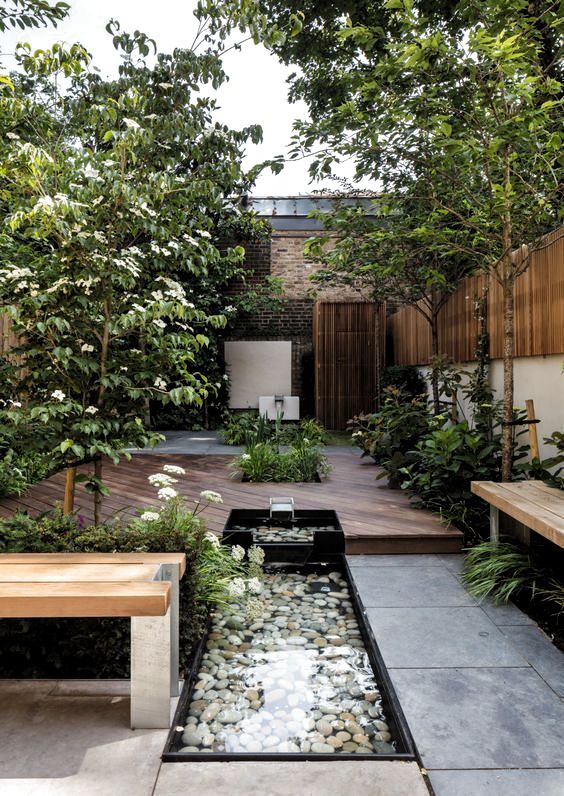 a beautiful summer terrace with trees, blooms and greenery, a fountain with pebbles, a bench and a deck