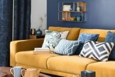 a chic living room with a navy wall, a yellow sofa and a rug, printed pillows, a cool gallery wall and wooden tables