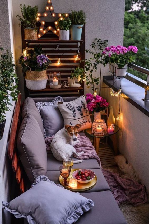 a cozy small balcony with a pallet wall and sofa, some potted blooms and greenery and candle lanterns