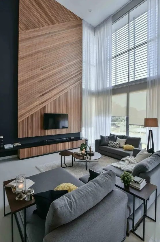 A gorgeous contemporary living room with a wood clad accent wall, a TV, grey furniture, side and coffee tables and double height windows
