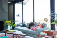a mid-century modern living room with a black brick fireplace, a light blue sofa with bright pillows, a colorful geo printed rug, potted greenery and wooden coffee tables