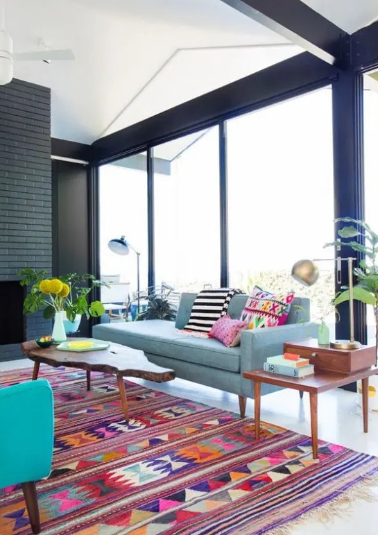A mid century modern living room with a black brick fireplace, a light blue sofa with bright pillows, a colorful geo printed rug, potted greenery and wooden coffee tables